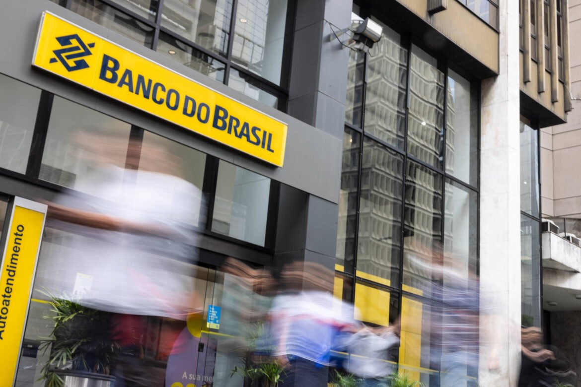 A imagem mostra a fachada de uma agência do Banco do Brasil. O logotipo do banco é visível em amarelo e azul, com o nome 'Banco do Brasil' em destaque. Há pessoas em movimento na frente da agência, sugerindo um ambiente movimentado. A estrutura do prédio é moderna, com grandes janelas refletindo o ambiente urbano ao redor. Fachada da agência do Banco do Brasil na avenida Paulista, no centro de São Paulo - Pedro Affonso - 30.jan.25/Folhapress