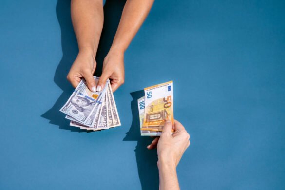 A imagem mostra duas mãos em um fundo azul. Uma mão está segurando várias notas de dólar, enquanto a outra mão está segurando uma nota de cinquenta euros. As mãos estão se aproximando, sugerindo uma troca de dinheiro. Brasileiros ainda têm valores a receber pelo Banco Central - Gabriel Cabral - 05.out.23/Folhapress